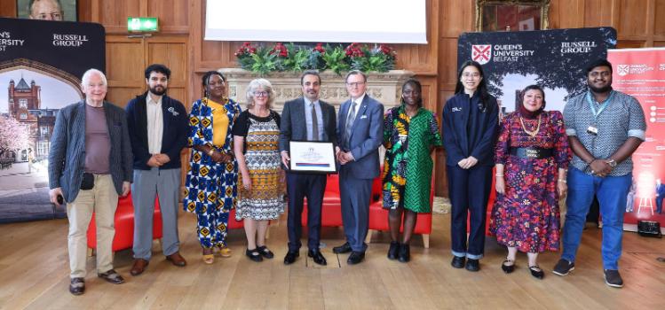 10 QUB Staff and student's union members standing in front of a decorative fireplace in a wood panled hall, with the two people in the middle holding the University of Sanctuary certificate